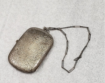 May include: An antique silver-toned rectangular purse with ornate floral and scrollwork engraving. The purse features a decorative monogram and a delicate chain strap. The clasp is closed. The overall design is vintage and elegant.