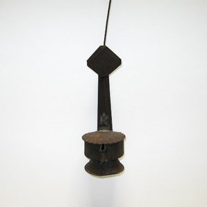 May include: A vintage, dark metal candle holder with a diamond-shaped top, a long stem, and a circular base. The candle holder is suspended by a thin wire against a white background. The metal has a rustic, aged appearance.