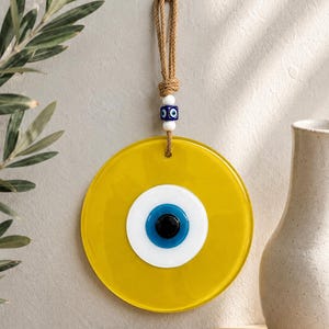 May include: A circular yellow glass wall hanging featuring a central eye motif with white, blue, and black concentric circles. The piece is suspended by a tan rope adorned with a small blue and white evil eye bead and two white spacer beads.