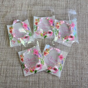 Può includere: Cinque piccoli sacchetti di plastica trasparente, ognuno contenente un braccialetto di perline rosa e bianco con un motivo floreale e le parole "Grazie".