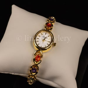 Pu&ograve; includere: Un orologio da polso dorato con quadrante ovale e bianco. Il cinturino dell'orologio presenta pietre preziose ovali colorate in rosso, viola e verde. L'orologio &egrave; esposto su un cuscino bianco.