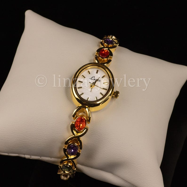 Pu&ograve; includere: Un orologio da polso dorato con quadrante ovale e bianco. Il cinturino dell'orologio presenta pietre preziose ovali colorate in rosso, viola e verde. L'orologio &egrave; esposto su un cuscino bianco.