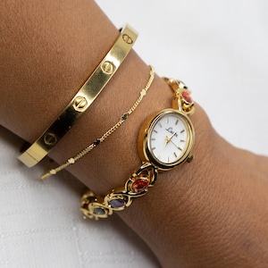 Pu&ograve; includere: Un orologio dorato con quadrante bianco e pietre preziose colorate, abbinato a un braccialetto dorato e a un delicato braccialetto a catena dorata. L'orologio presenta il nome del marchio "Lucian" sul quadrante. I braccialetti e l'orologio sono indossati su un polso.