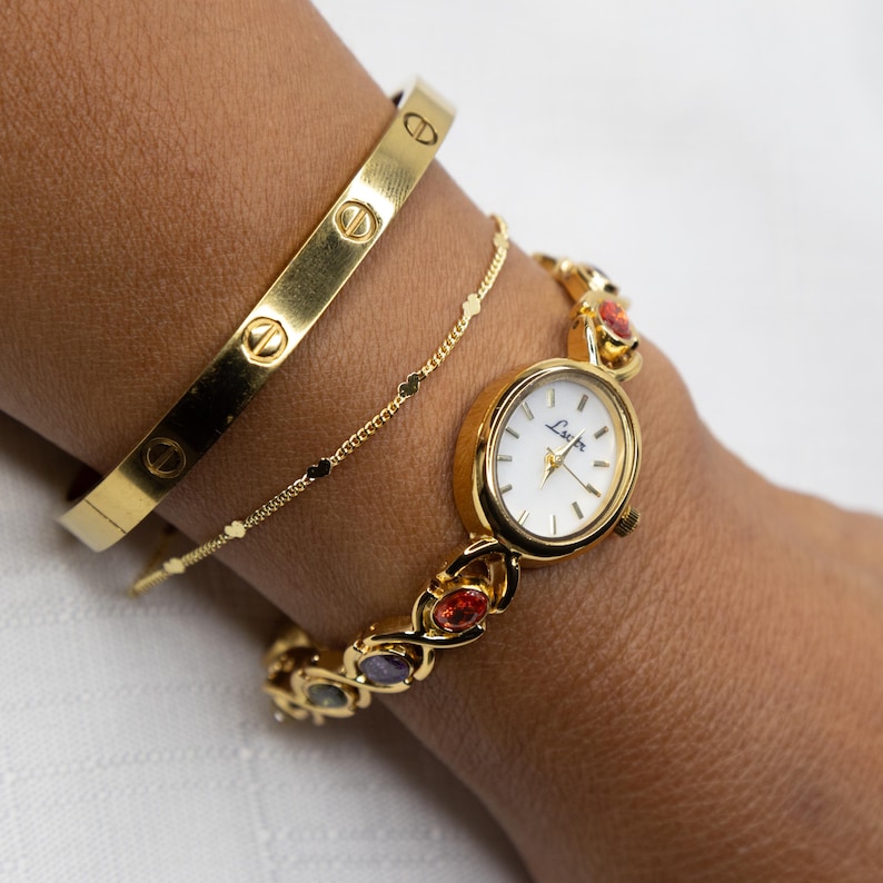 Pu&ograve; includere: Un orologio dorato con quadrante bianco e pietre preziose colorate, abbinato a un braccialetto dorato e a un delicato braccialetto a catena dorata. L'orologio presenta il nome del marchio "Lucian" sul quadrante. I braccialetti e l'orologio sono indossati su un polso.