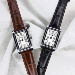 Könnte beinhalten: Zwei rechteckige Armbanduhren mit silbernen Gehäusen und weißen Zifferblättern, mit schwarzen römischen Ziffern und Zeigern. Eine Uhr hat ein braunes Lederarmband, die andere ein schwarzes Lederarmband. Das Zifferblatt zeigt "ORSGA".