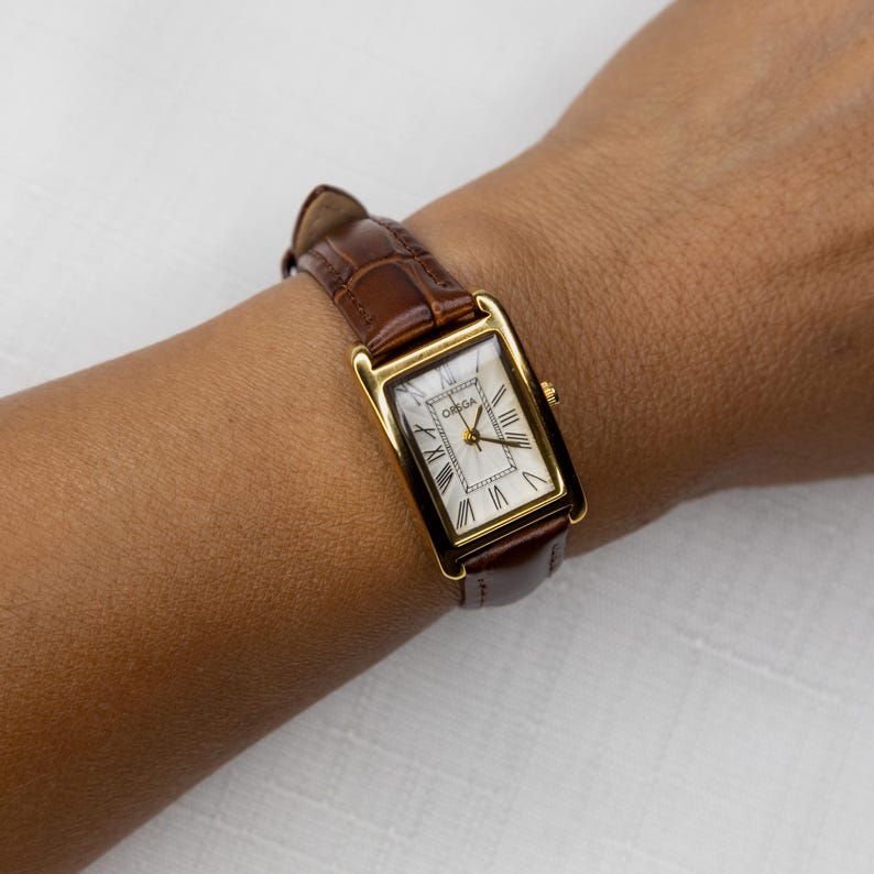 May include: A gold rectangular wristwatch with a brown leather band. The watch face is white with black Roman numerals and gold hands. The watch is marked "OMEGA".