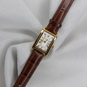 May include: A gold rectangular wristwatch with a white face and Roman numerals. The watch has a brown leather band. The watch face has the brand name "ORSGA" printed on it.