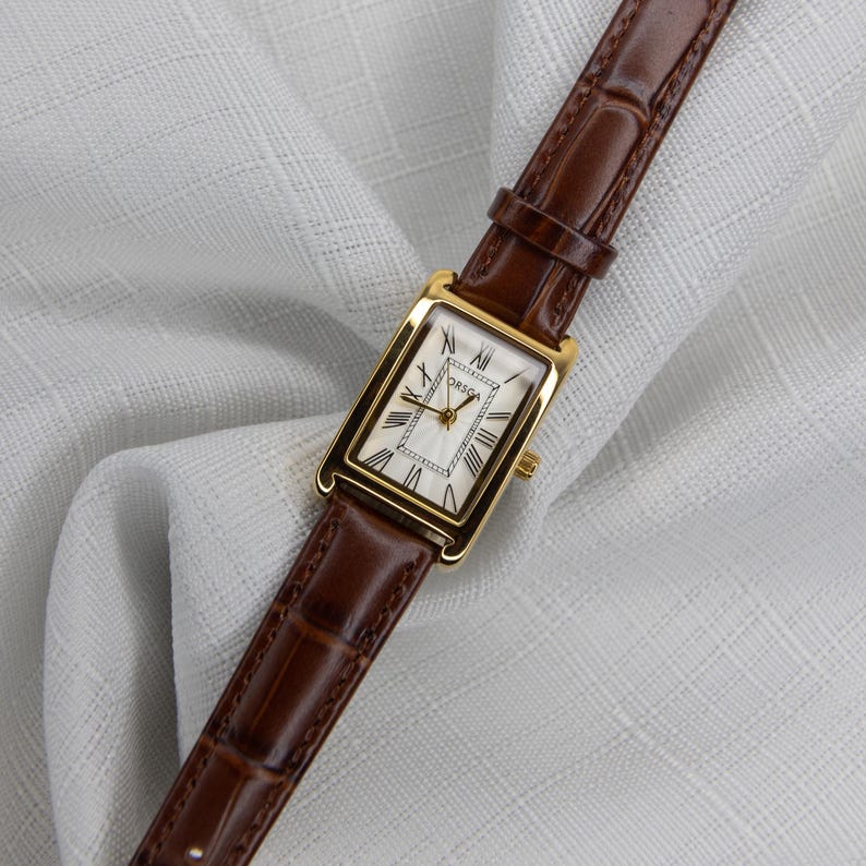 May include: A gold rectangular wristwatch with a white face and Roman numerals. The watch has a brown leather band. The watch face has the brand name "ORSGA" printed on it.