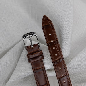 May include: Close-up of a brown leather watch strap with a silver buckle. The strap has a textured, crocodile-like pattern. The buckle is engraved with the word "ORSGA".