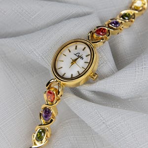 Pu&ograve; includere: Orologio da polso dorato con quadrante bianco e marchio "Lyter". Il cinturino dell'orologio presenta una serie di pietre preziose colorate rosse, arancioni, viola, verdi e gialle. L'orologio &egrave; esposto su un tessuto bianco.
