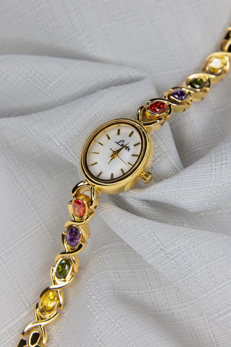 Pu&ograve; includere: Orologio da polso dorato con quadrante bianco e marchio "Lyter". Il cinturino dell'orologio presenta una serie di pietre preziose colorate rosse, arancioni, viola, verdi e gialle. L'orologio &egrave; esposto su un tessuto bianco.