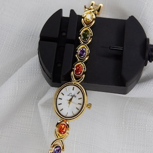 Pu&ograve; includere: Orologio da polso dorato con quadrante bianco e marchio "Lsofter". Il cinturino presenta una serie di pietre preziose colorate in giallo, verde, viola e rosso. L'orologio &egrave; esposto su uno strumento nero per la riparazione di orologi.