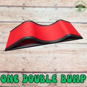May include: A red, double-bump shaped object with black trim, set against a wood-grain background. The text "ONE DOUBLE BUMP" is displayed in green, below the object. A logo for "Flying Saucer Printing" is in the upper right corner.