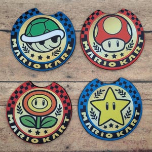 May include: Four round, colorful Mario Kart themed car coasters. Each coaster features a different character from the game: a green turtle shell, a red mushroom, a yellow star, and a yellow flower.