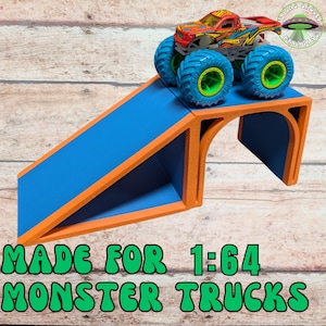 May include: A blue and orange ramp for 1:64 monster trucks. The ramp has a blue surface with orange edges and supports. A red, yellow, and blue monster truck with large blue tires is on top of the ramp. The text "MADE FOR 1:64 MONSTER TRUCKS" is at the bottom.