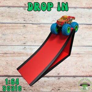 Peut inclure: Une rampe rouge avec des bordures noires supporte un monster truck color&eacute; avec des roues bleues et vertes. Les mots "DROP IN" sont en vert en haut, et "1:64 scale" en bas. Un logo Flying Saucer Printing est en bas &agrave; droite.