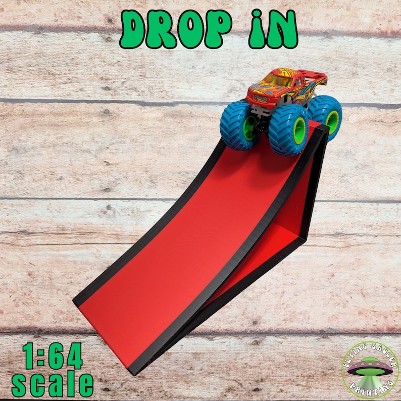 Peut inclure: Une rampe rouge avec des bordures noires supporte un monster truck color&eacute; avec des roues bleues et vertes. Les mots "DROP IN" sont en vert en haut, et "1:64 scale" en bas. Un logo Flying Saucer Printing est en bas &agrave; droite.