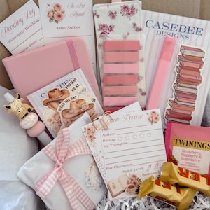 May include: A curated book lover's gift box containing a pink notebook, a floral fabric bookmark with sticky tabs, a reading log, a book review card, a pen with a highland cow topper, a stack of books sticker, a Twinings tea bag, and two candy bars.