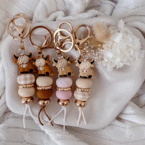 May include: Four keychains featuring Highland cow charms attached to gold-toned metal rings. Each keychain includes a sequence of beads in various textures and patterns, including solid beige, white gingham, pink, and brown, finished with short white or brown cords.