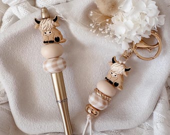 May include: A gold-toned pen and a matching keychain, both featuring a silicone highland cow charm and beige-toned beads, including one with a white and beige gingham pattern. The keychain includes a gold metal ring and a small white tassel.