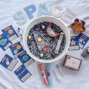 Pode incluir: Um kit sensorial com tema espacial contendo uma tigela branca cheia de enchimento preto e branco, um foguete de madeira, uma lua crescente, estrelas, um tubo de ensaio, figuras de astronautas, cartões de fatos espaciais e pequenos frascos.