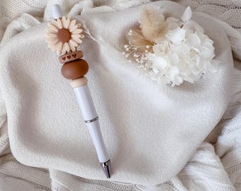 May include: A white ballpoint pen featuring a decorative topper with a beige sunflower, a small brown bead with three engraved hearts, and a larger smooth brown bead. The pen has a silver-toned metal tip and a matching silver-toned center ring.