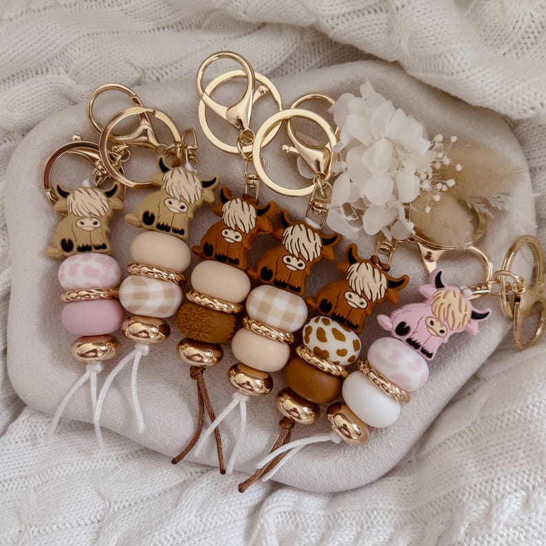 May include: A collection of five keychains featuring cartoon highland cow charms attached to gold-toned metal rings. Each keychain includes a sequence of round beads in various shades of pink, white, tan, and brown, with some beads displaying checkered or spotted patterns.