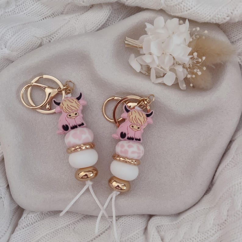 May include: Two keychains featuring a pink highland cow charm attached to a gold-tone metal ring. Each keychain includes a sequence of beads: one pink and white patterned bead, one white bead, and two gold-tone metallic beads, finished with a white cord.