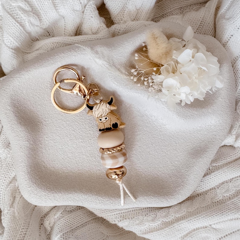 May include: A keychain featuring a gold-toned metal ring and clasp attached to a silicone highland cow charm. The charm is followed by a series of beads in shades of beige and cream, including a smooth round bead, a textured bead, and a metallic bead.