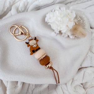 May include: A keychain featuring a brown Highland cow silicone charm, a smooth cream bead, a textured brown bead, and a metallic gold bead. The charm is attached to a gold-toned metal split ring and lobster clasp, finished with a thin brown cord.