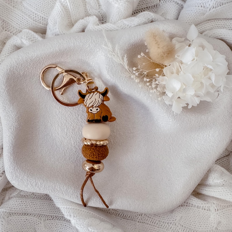 May include: A keychain featuring a brown Highland cow charm, a smooth cream bead, a gold-toned textured spacer, a dark brown textured bead, and a gold-toned metallic bead, all attached to a gold-toned metal split ring and lobster clasp.