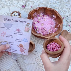 May include: A wooden bowl containing a pink bubbly liquid with flower petals, a small wooden cup filled with pink shimmering rocks, and an instruction card titled Fairy Glow Potion with illustrations of a bowl, rocks, petals, and a fairy.