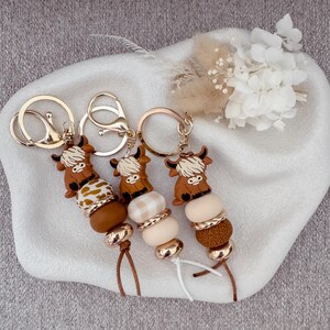 May include: Three keychains featuring brown Highland cow charms attached to silver-tone metal rings. Each keychain includes a series of beads in various shades of brown, cream, and white, with some featuring patterns and metallic accents, finished with a short cord.