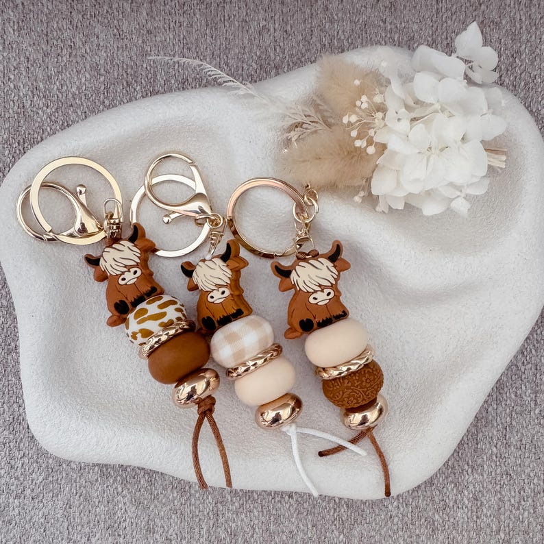 May include: Three keychains featuring brown Highland cow charms attached to silver-tone metal rings. Each keychain includes a series of beads in various shades of brown, cream, and white, with some featuring patterns and metallic accents, finished with a short cord.
