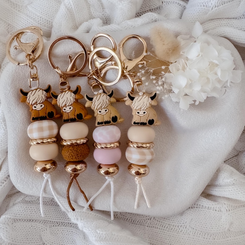 May include: Four keychains featuring silicone Highland cow charms in brown and cream tones. Each charm is attached to a gold-toned metal ring and hangs above a stack of round beads in various textures, including gingham patterns, solid colors, and metallic gold accents.