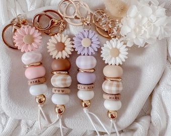 May include: Four keychains featuring silicone flower charms in pink, beige, lavender, and white. Each includes round silicone and wooden beads, some with gingham patterns or the text MAMA, KIND, and heart icons, attached to gold metal rings.