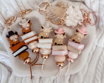 May include: Four keychains featuring silicone highland cow figures wearing cowboy hats, each perched atop stacked silicone book beads and round beads in various shades of brown, white, and pink, finished with rose gold metal lobster clasps.