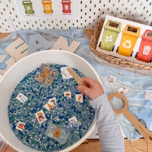 May include: A white sensory bin filled with blue and green pebbles, containing small square cards depicting various waste items, a clear resin block with a crab, and a clear resin block with a starfish. A wooden scoop and a net are nearby.