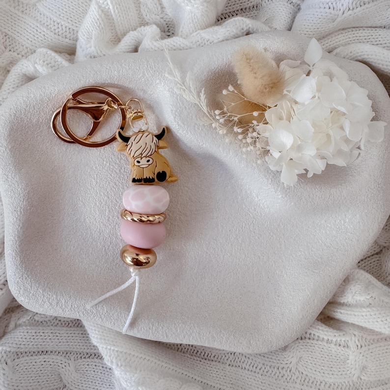 May include: A keychain featuring a tan Highland cow charm with black horns, attached to a gold-toned metal ring. The charm is followed by a sequence of beads including a pink and white patterned bead, a gold-toned ring, a solid pink bead, and a gold-toned bead.