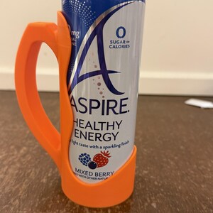 Op de afbeelding: Een oranje plastic blikhouder met een handvat, met een blikje Aspire Healthy Energy. Het blikje is blauw en wit met de woorden "Mixed Berry" en "0 Sugar or Calories" erop gedrukt.