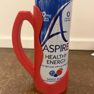 Op de afbeelding: Een rode plastic handgreep omhult een blauw en wit blikje van de drank "ASPIRE HEALTHY ENERGY". Op het blikje staat de tekst "0mg NATUURLIJKE CAFE&Iuml;NE" en "0 SUIKER CALORIE&Euml;N". Het blikje is op smaak gebracht met "MIXED BERRY".