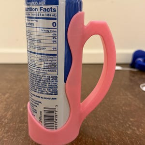 Op de afbeelding: Een roze 3D-geprinte blikhouder met een handvat, ontworpen voor een standaard 355 ml blikje. De houder omsluit het blikje en biedt een grip. Het blikje heeft een blauw en wit etiket met voedingsfeiten.