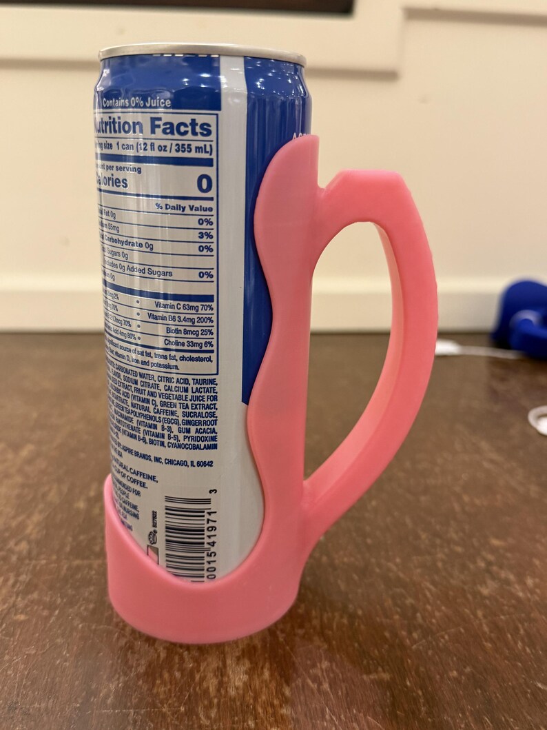 Op de afbeelding: Een roze 3D-geprinte blikhouder met een handvat, ontworpen voor een standaard 355 ml blikje. De houder omsluit het blikje en biedt een grip. Het blikje heeft een blauw en wit etiket met voedingsfeiten.