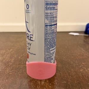 Op de afbeelding: Een zilver-blauwe Aspire Healthy Energy drankblik met een roze siliconen basis. De blik heeft voedingsfeiten en de woorden "RE" en "Healthy Energy" erop gedrukt. De basis is lichtroze en heeft een logo.