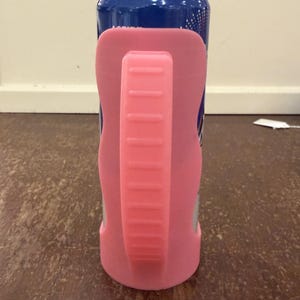 Op de afbeelding: Een blauw en zilver drankblikje met een roze handvat en houder. De roze houder omsluit het blikje en biedt een grip. Het handvat is effen roze met horizontale ribbels.