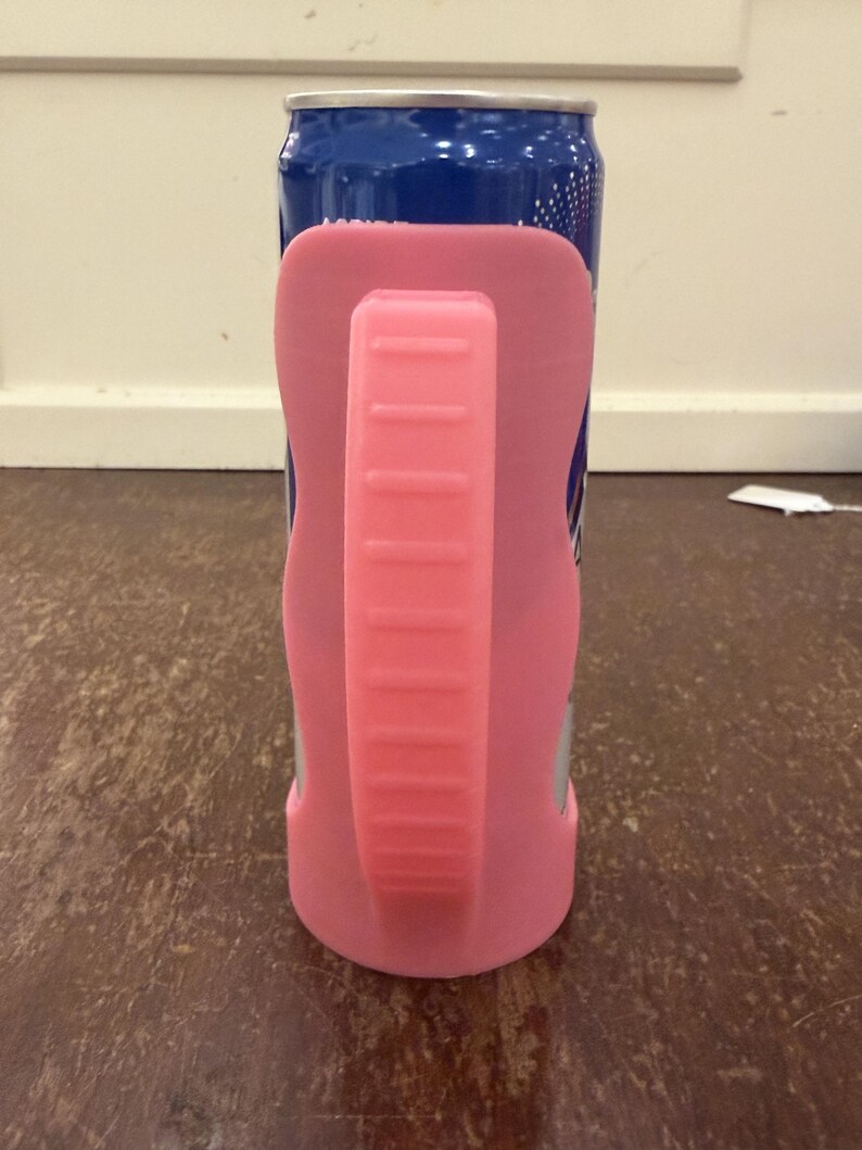 Op de afbeelding: Een blauw en zilver drankblikje met een roze handvat en houder. De roze houder omsluit het blikje en biedt een grip. Het handvat is effen roze met horizontale ribbels.