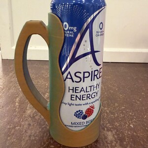 Op de afbeelding: Een blikje Aspire Healthy Energy-drank in een op maat gemaakte houder. Het blikje is wit en blauw met de tekst "Aspire Healthy Energy" en "Mixed Berry". De houder is groen en goudkleurig met het woord "Jungle" onderaan.