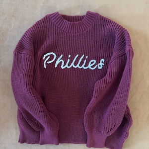 Könnte beinhalten: Ein weinroter Strickpullover mit dem Wort "Phillies" in Hellblau auf der Brust gestickt. Der Pullover hat einen gerippten Rundhalsausschnitt, lange Ärmel und eine lockere Passform. Die Ärmel haben eine leichte Ballonform.
