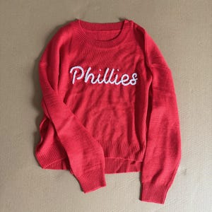 May include: A red knit sweater with the word "Phillies" embroidered in white across the chest. The sweater has a crew neck and ribbed cuffs and hem. The sleeves are long and the overall style is casual.