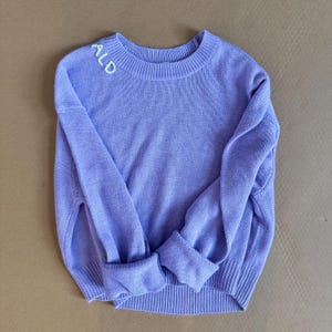 May include: A lavender knitted sweater with the word "ALD" embroidered on the neckline.
